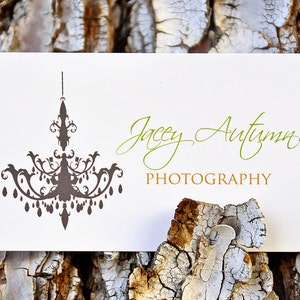 May include: A rectangular white business card with rounded corners featuring a dark brown chandelier graphic on the left side. The text Jacey Autumn is printed in green script, and the word PHOTOGRAPHY is printed in gold block letters below it.