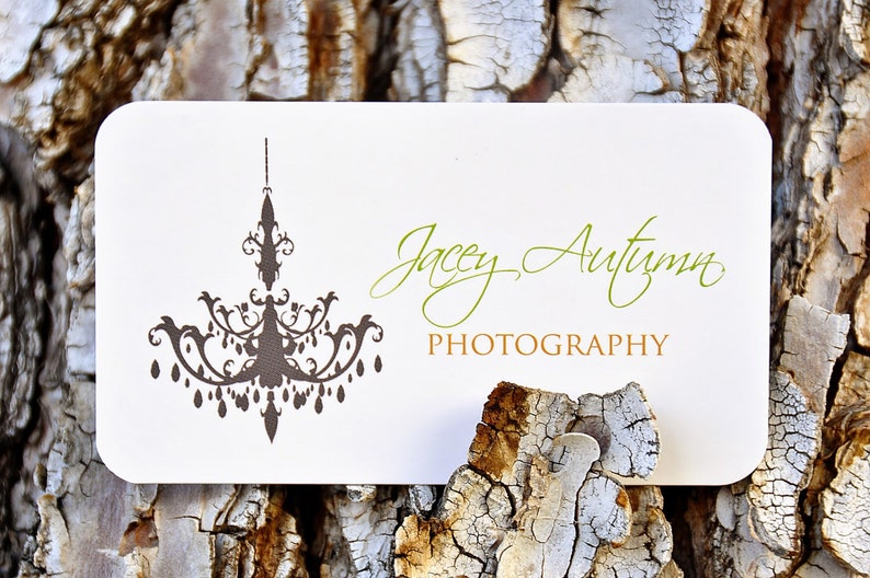 May include: A rectangular white business card with rounded corners featuring a dark brown chandelier graphic on the left side. The text Jacey Autumn is printed in green script, and the word PHOTOGRAPHY is printed in gold block letters below it.