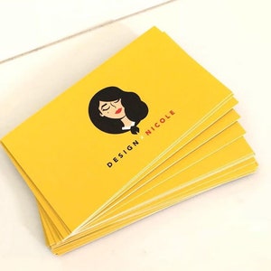 May include: A stack of rectangular business cards in a bright yellow color. Each card features a black circular graphic of a stylized face with dark hair and red lips, accompanied by the black and red text that reads DESIGN x NICOLE.