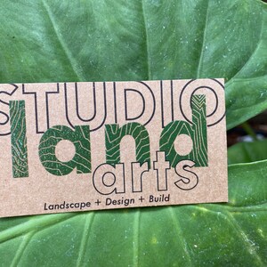 May include: A business card with the words "STUDIO LAND arts" in black and green. The card is on a large green leaf. The text "Landscape + Design + Build" is below the main text.