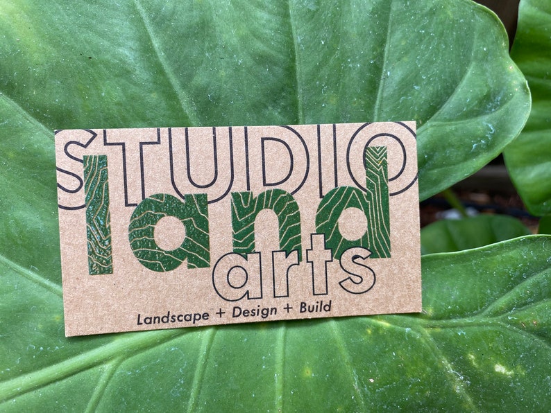 May include: A business card with the words "STUDIO LAND arts" in black and green. The card is on a large green leaf. The text "Landscape + Design + Build" is below the main text.