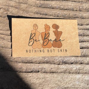 May include: A rectangular tan card featuring a black line art illustration of three figures from the back. The text "Be Bare" is written in a cursive script above the words "NOTHING BUT SKIN" printed in a clean, sans-serif black font.