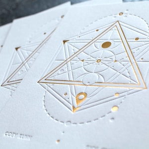 May include: A set of white rectangular cards featuring an embossed geometric design with gold foil accents. The central motif consists of a large triangle, intersecting lines, and circular elements. The text COEY KUNN is embossed at the bottom.