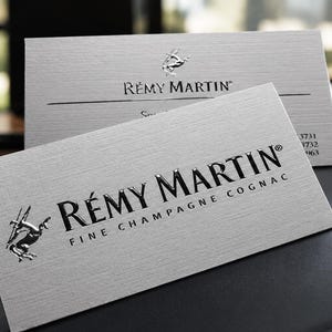 K&ouml;nnte beinhalten: Zwei rechteckige wei&szlig;e Visitenkarten mit strukturierter Oberfl&auml;che. Die Karten zeigen den schwarzen gepr&auml;gten Text R&Eacute;MY MARTIN und die Worte FINE CHAMPAGNE COGNAC, begleitet von einem silbernen metallischen Zentaur-Logo links.