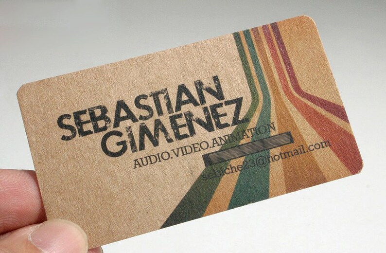 May include: A rectangular business card made of light brown textured paper. The card features the black text SEBASTIAN GIMENEZ, AUDIO.VIDEO.ANIMATION, and sebiche23@hotmail.com. Diagonal stripes in green, tan, and red extend across the right side.
