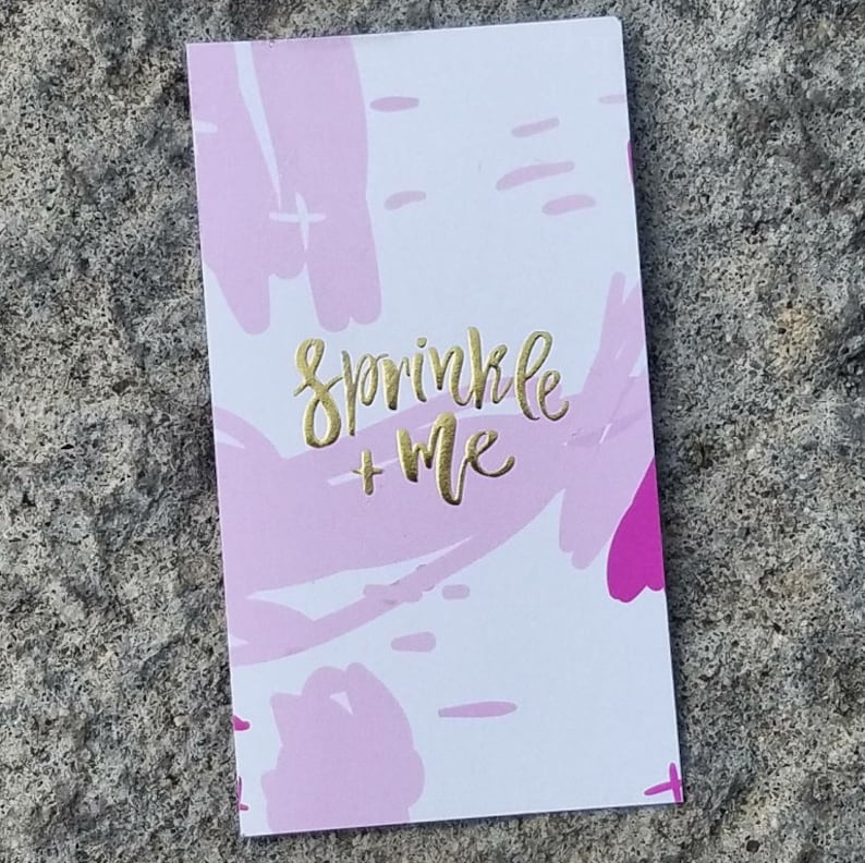 Puede incluir: Una tarjeta rectangular blanca con un patr&oacute;n de pinceladas abstractas en rosa y magenta. El texto "sprinkle + me" est&aacute; impreso en el centro con una fuente cursiva de color dorado met&aacute;lico. La tarjeta tiene un acabado liso y bordes rectos.