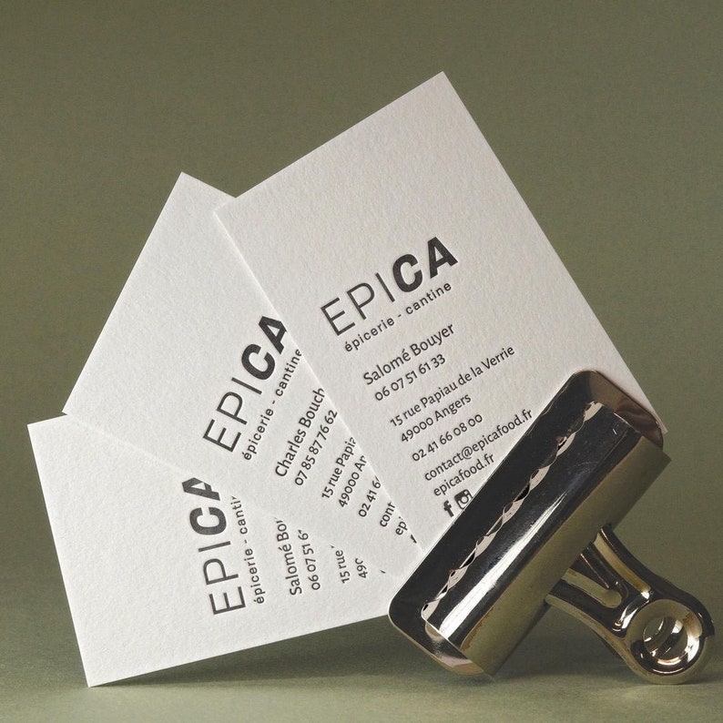 以下が含まれることがあります： EPICA &eacute;picerie - cantineという黒い文字と連絡先、住所が記載された3枚の白い長方形の名刺。名刺は右下の光沢のある銀色の金属製バインダークリップでまとめられています。