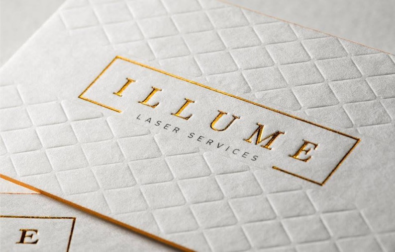May include: A white business card with a textured diamond pattern embossed across the surface. The card features the gold foil stamped text ILLUME and the smaller black text LASER SERVICES enclosed within a thin gold rectangular border.