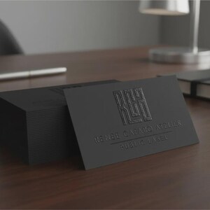 May include: A stack of rectangular black business cards with a matte finish. The top card features an embossed logo with the letters RCH and the text RENEE CABARO ATELIER & PUBLIC LABEL printed in a subtle, dark, and raised texture on the surface.
