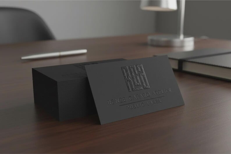 May include: A stack of rectangular black business cards with a matte finish. The top card features an embossed logo with the letters RCH and the text RENEE CABARO ATELIER & PUBLIC LABEL printed in a subtle, dark, and raised texture on the surface.