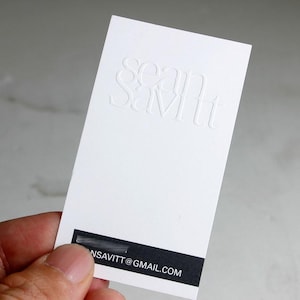 May include: A white business card with the embossed name "sean savitt" at the top. The bottom of the card has a black stripe with an email address. The card is held by a person.