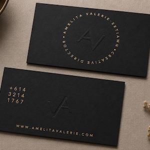 May include: Two rectangular black business cards with gold foil lettering. The top card features a circular design with the text AMELITA VALERIE SETIAWN CREATIVE DIRECTOR and the initials AV. The bottom card displays the phone number +614 3214 1767, the initials VA, and the website WWW.AMELITAVALERIE.COM.