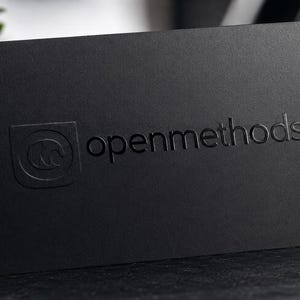 May include: A rectangular black business card featuring the text openmethods embossed on the front surface. To the left of the text is a square logo containing a stylized letter M. The card has a matte finish and a clean, professional appearance.