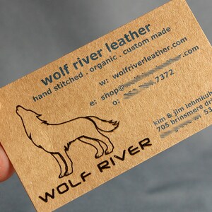 May include: A business card with the text "wolf river leather" and a howling wolf illustration. The card is a natural brown color with black text and a logo. The card also includes contact information.