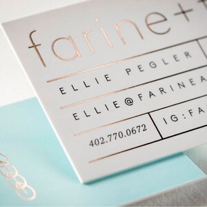 May include: A white business card with rose gold foil lettering reading farine + four, Ellie Pegler, and contact details, layered over a light blue card featuring a rose gold wheat stalk icon. The text includes an email address and phone number.