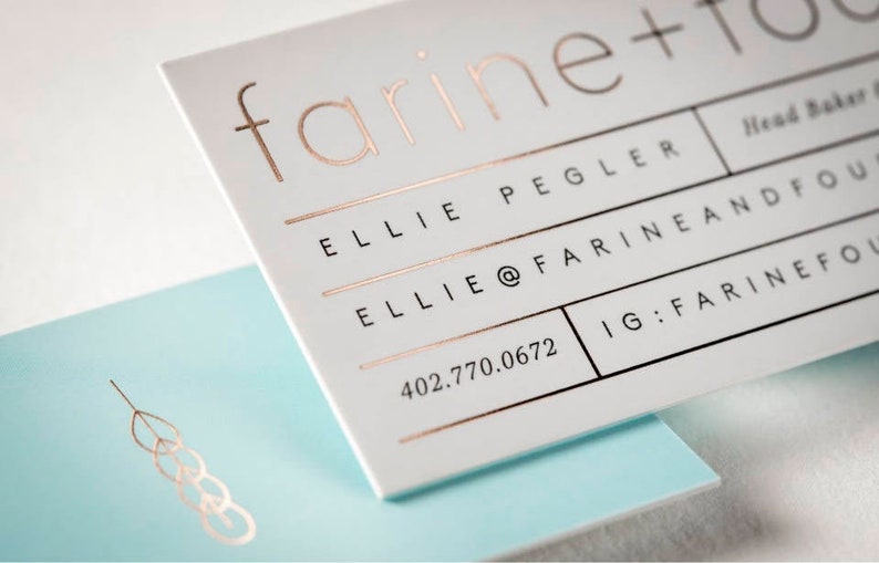 May include: A white business card with rose gold foil lettering reading farine + four, Ellie Pegler, and contact details, layered over a light blue card featuring a rose gold wheat stalk icon. The text includes an email address and phone number.
