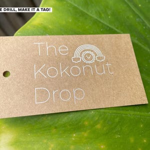 May include: A rectangular brown kraft paper tag with a pre-punched hole on the left side. The text "The Kokonut Drop" is printed in white, accompanied by a small white line-art graphic of a smiling sun positioned beneath a rainbow arch.