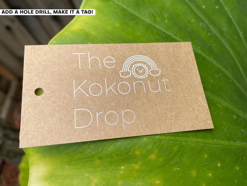 May include: A rectangular brown kraft paper tag with a pre-punched hole on the left side. The text "The Kokonut Drop" is printed in white, accompanied by a small white line-art graphic of a smiling sun positioned beneath a rainbow arch.