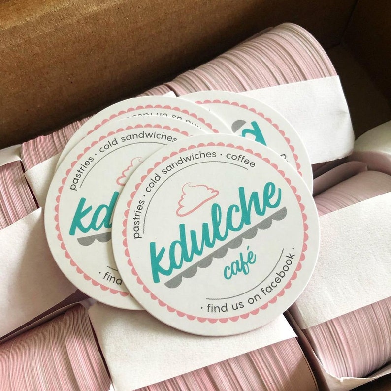 Puede incluir: Una pila de posavasos redondos de papel blanco con un borde festoneado rosa y el texto en color verde azulado kdulche caf&eacute;. El dise&ntilde;o incluye un peque&ntilde;o icono de pastel rosa, las palabras pastries, cold sandwiches, coffee y find us on facebook.