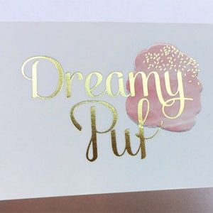 Puede incluir: Una tarjeta de visita rectangular blanca con el texto Dreamy Puff en l&aacute;mina dorada y tipograf&iacute;a cursiva. A la derecha del texto hay un gr&aacute;fico rosa suave estilo acuarela con peque&ntilde;as motas blancas que parecen chispas de az&uacute;car.