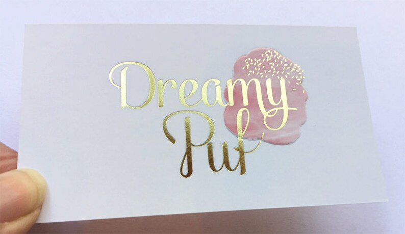 Puede incluir: Una tarjeta de visita rectangular blanca con el texto Dreamy Puff en l&aacute;mina dorada y tipograf&iacute;a cursiva. A la derecha del texto hay un gr&aacute;fico rosa suave estilo acuarela con peque&ntilde;as motas blancas que parecen chispas de az&uacute;car.