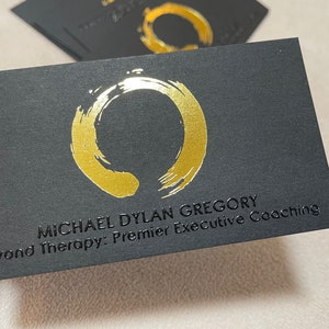 May include: A rectangular black business card featuring a metallic gold brushstroke circle logo. The text MICHAEL DYLAN GREGORY and Beyond Therapy: Premier Executive Coaching is embossed in black on the card surface. Additional cards are visible.