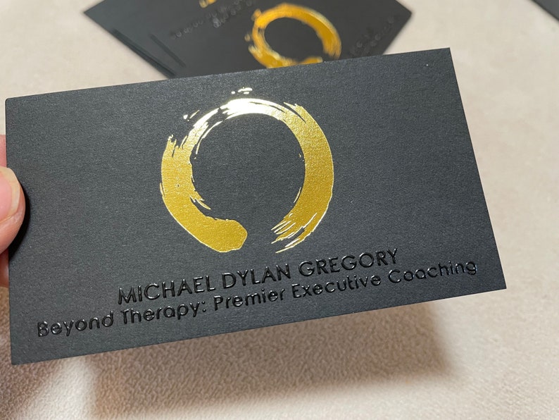 May include: A rectangular black business card featuring a metallic gold brushstroke circle logo. The text MICHAEL DYLAN GREGORY and Beyond Therapy: Premier Executive Coaching is embossed in black on the card surface. Additional cards are visible.