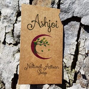 May include: A rectangular brown kraft paper tag featuring the black text Ashjoi at the top and Natural Artisan Soap at the bottom. In the centre is a maroon crescent moon graphic containing a tree with green leaves and small falling leaves.