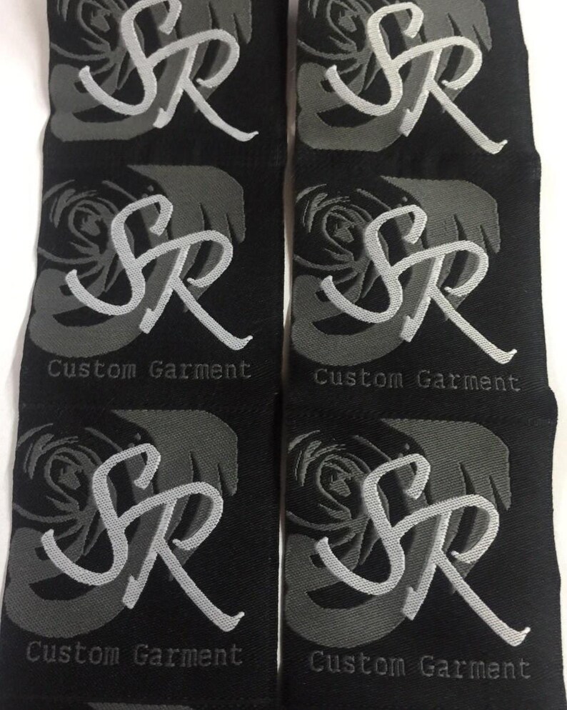 May include: Six black fabric labels arranged in two columns. Each label features a dark gray rose graphic, the light gray embroidered letters SR, and the text Custom Garment printed in a small, simple font at the bottom of each individual label.