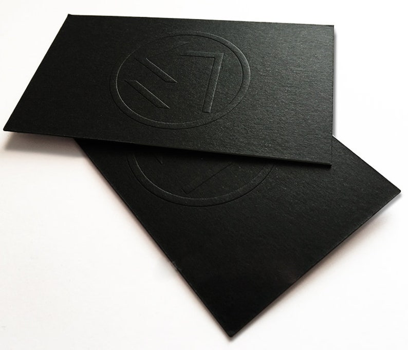 May include: Two rectangular black business cards stacked on top of each other. Each card features a circular embossed logo in the center, consisting of a geometric shape resembling a stylized letter or symbol enclosed within a thin circular border.