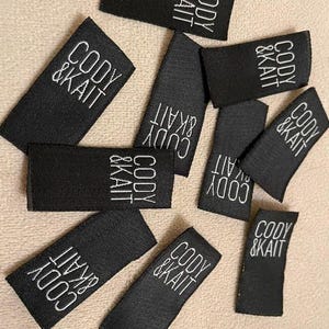 May include: A collection of rectangular black fabric labels featuring the white embroidered text CODY & KAIT. These small, woven garment tags are arranged in a scattered pattern, each displaying the brand name in a clean, sans-serif font.