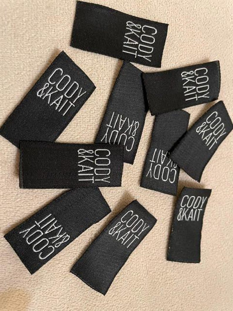 May include: A collection of rectangular black fabric labels featuring the white embroidered text CODY & KAIT. These small, woven garment tags are arranged in a scattered pattern, each displaying the brand name in a clean, sans-serif font.