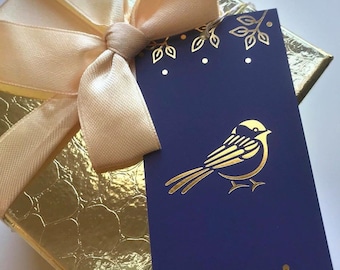 Puede incluir: Una caja de regalo dorada con superficie texturizada y un lazo de satén beige claro. Sujeta a la cinta hay una etiqueta rectangular azul oscuro con una ilustración en lámina dorada de un pájaro pequeño rodeado de hojas y puntos.
