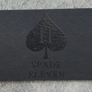 May include: A rectangular black business card featuring a textured spade symbol at the center with the number eleven integrated into the design. Below the spade, the text SPADE ELEVEN is embossed in a clean, sans-serif font on the matte surface.