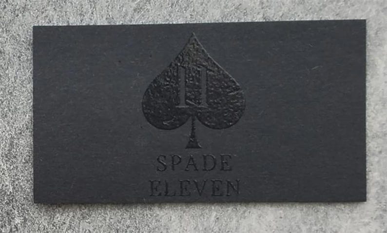 May include: A rectangular black business card featuring a textured spade symbol at the center with the number eleven integrated into the design. Below the spade, the text SPADE ELEVEN is embossed in a clean, sans-serif font on the matte surface.