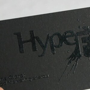 May include: A rectangular black business card featuring the embossed text HypeFX with a glossy ink splatter effect. The card includes the name Chris Ciardi, the title Head Marketing, and contact information printed in a subtle black font.