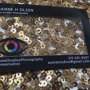 Puede incluir: Una tarjeta de visita rectangular negra con un centro transparente, que muestra el texto ROANNE H OLSEN, Aume Studios Photography, un icono de lente de c&aacute;mara en forma de ojo colorido, redes sociales, un n&uacute;mero de tel&eacute;fono y un correo electr&oacute;nico.