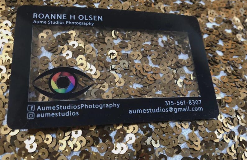 Puede incluir: Una tarjeta de visita rectangular negra con un centro transparente, que muestra el texto ROANNE H OLSEN, Aume Studios Photography, un icono de lente de c&aacute;mara en forma de ojo colorido, redes sociales, un n&uacute;mero de tel&eacute;fono y un correo electr&oacute;nico.