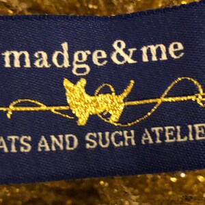 May include: A rectangular blue fabric clothing label featuring the white text madge & me, a gold silhouette of a dog, and the text HATS AND SUCH ATELIER in white. The label is positioned against a background of shimmering gold metallic fibers.