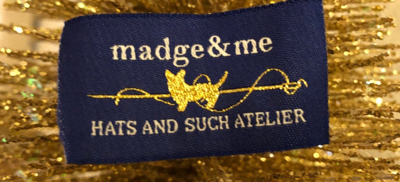 May include: A rectangular blue fabric clothing label featuring the white text madge & me, a gold silhouette of a dog, and the text HATS AND SUCH ATELIER in white. The label is positioned against a background of shimmering gold metallic fibers.