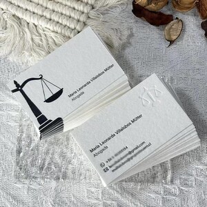 May include: Two stacks of white rectangular business cards. The top card of the left stack features a black graphic of a scale of justice and the text Maria Leonarda Villalobos M&uuml;tter Abogada. The right stack shows an embossed scale of justice.