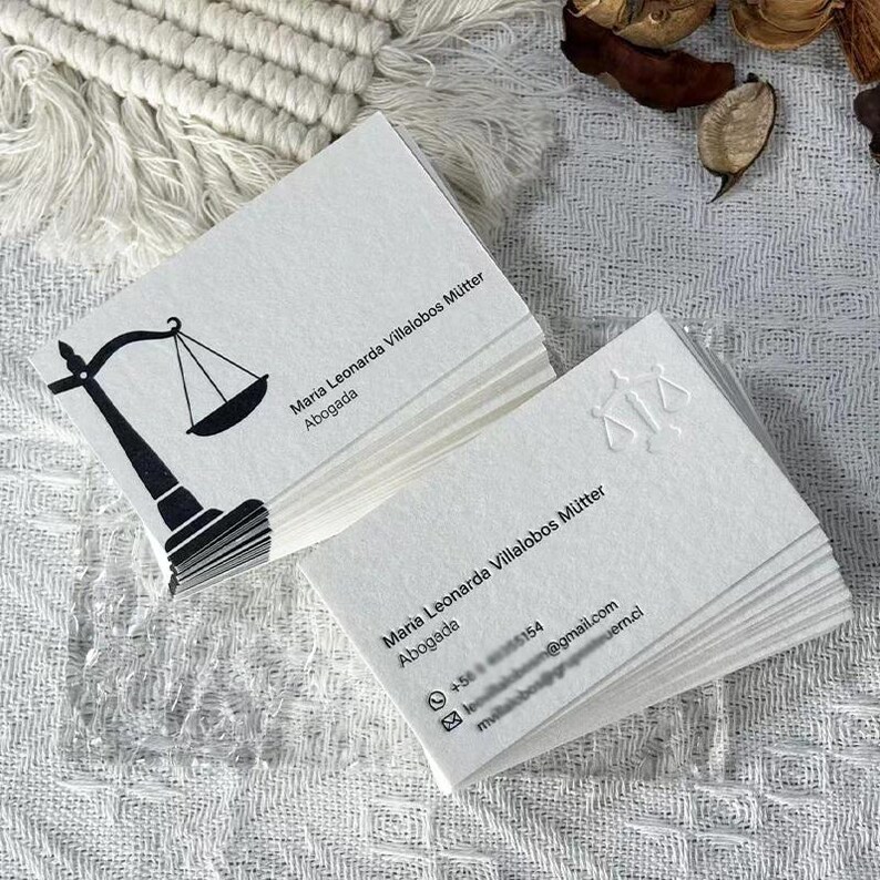 May include: Two stacks of white rectangular business cards. The top card of the left stack features a black graphic of a scale of justice and the text Maria Leonarda Villalobos M&uuml;tter Abogada. The right stack shows an embossed scale of justice.