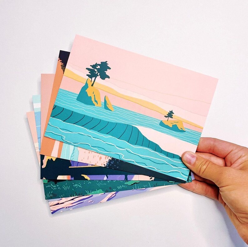 May include: A stack of rectangular postcards featuring colorful graphic illustrations of landscapes. The top postcard displays a coastal scene with turquoise water, yellow rock formations, and small trees under a pale pink sky. Other cards show varied nature scenes.
