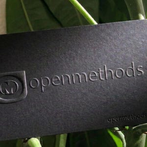 May include: A rectangular black business card featuring a debossed logo of the letter M inside a shield shape, the text openmethods in a sans-serif font, and the website openmethods.com printed in the bottom right corner in a dark finish.