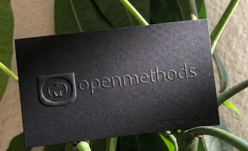May include: A rectangular black business card featuring a debossed logo of the letter M inside a shield shape, the text openmethods in a sans-serif font, and the website openmethods.com printed in the bottom right corner in a dark finish.
