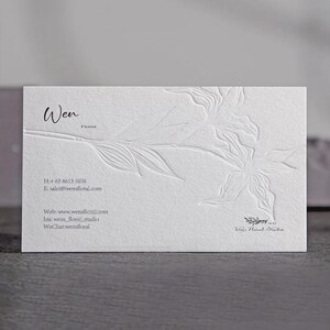 May include: A rectangular white business card featuring a blind embossed floral design on the right side. The text reads Wen Florist, along with contact details including a phone number, email address, website, Instagram, and WeChat handle.