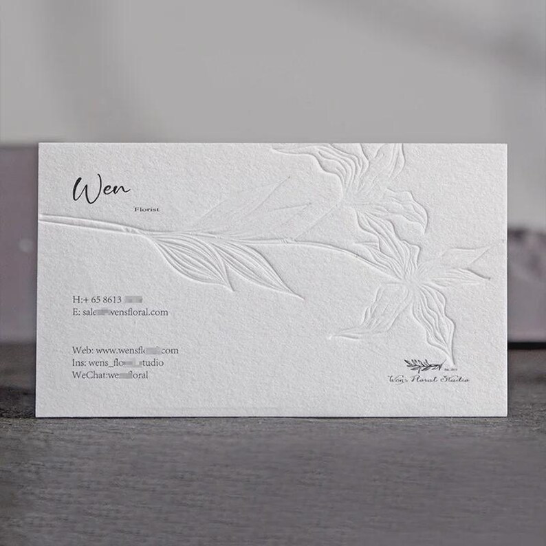 May include: A rectangular white business card featuring a blind embossed floral design on the right side. The text reads Wen Florist, along with contact details including a phone number, email address, website, Instagram, and WeChat handle.