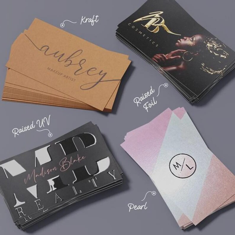 May include: Four stacks of rectangular business cards featuring different finishes. One stack is brown kraft paper with aubrey makeup artist text. One is black with gold raised foil and a portrait. One is black with raised UV text. One is pearl paper with M/L logo.