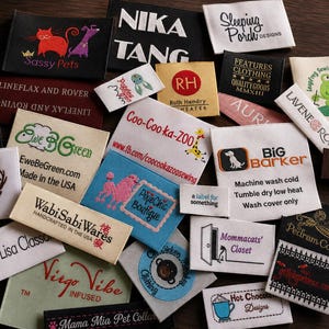 May include: A collection of various rectangular fabric clothing labels featuring diverse brand names, logos, and care instructions. The labels are printed or embroidered in multiple colors including black, white, blue, red, and green on different fabric textures.