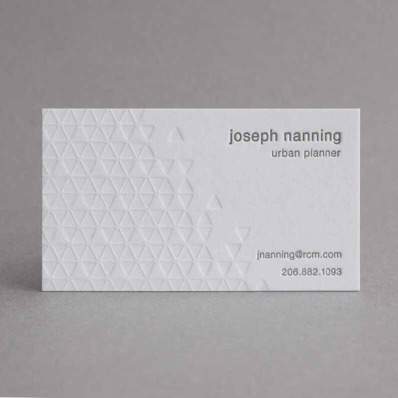 May include: A white rectangular business card featuring a debossed geometric triangle pattern on the left side. The text reads joseph nanning, urban planner, jnanning@rcm.com, and 206.882.1093 in a clean, dark gray sans-serif typeface.